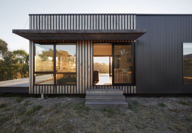 Exterior view of a compact modern dwelling with dark vertical cladding, a projecting timber-framed roof canopy, upper-level terrace balustrade, and large glazed openings. Warm interior light and furnishings are visible through the windows and doorway, with a small timber stair leading to the entrance. The building sits in a natural, lightly vegetated setting.
