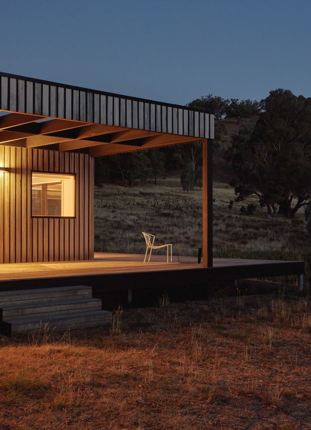 A compact modern cabin set in a natural bushland landscape at dusk. The building features dark vertical timber cladding, a flat roof with a projecting timber-framed covered deck, exposed roof beams, warm interior lighting, and broad open views toward the surrounding trees and dry grassland. A simple chair sits on the timber deck.