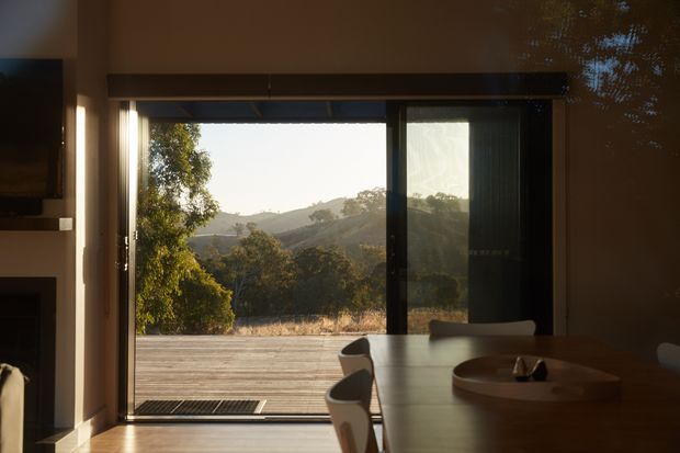 A dimly lit contemporary dining area viewed from inside, with a long dark dining table and chairs positioned beside full-height sliding glass doors. The doors open onto a timber deck, framing a sunlit rural landscape of trees and rolling hills. A television and built-in cabinetry are partially visible at left.
