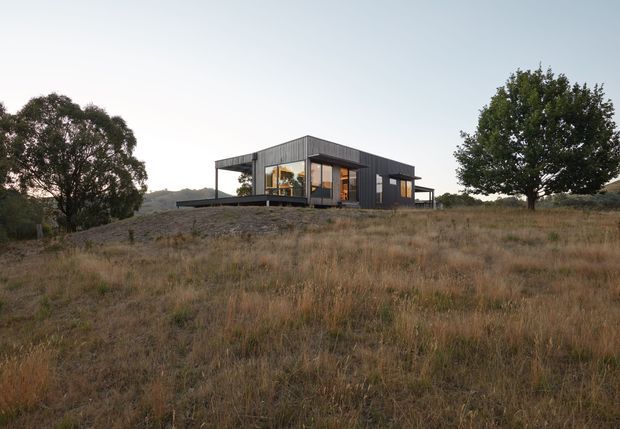 A compact contemporary single-storey house set in a dry rural landscape. The building has a strong rectangular form, dark vertical cladding, extensive floor-to-ceiling glazing, and broad covered decks or overhangs. Mature trees and rolling terrain surround the elevated-looking dwelling.
