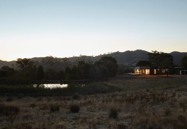 A small, low-profile contemporary dwelling sits in a dry rural landscape at dusk, with warmly illuminated floor-to-ceiling glazing and dark exterior walls. A reflective pond, scattered trees, rolling hills, and muted grasses surround the isolated home.