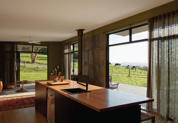 Warm, open-plan kitchen interior with a large timber island and integrated black sink, set against dark cabinetry and a textured stone or concrete feature wall. Full-height glazing and sheer curtains open to a green rural landscape with grazing animals, while patterned rugs and earthy finishes add warmth.