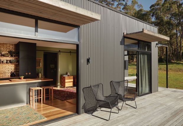 Contemporary single-storey home with a dark vertically ribbed metal-clad facade, broad timber-lined roof overhangs, and extensive floor-to-ceiling glazing opening onto a spacious timber deck. The open living and kitchen area is visible through the large sliding doors, with outdoor wire-frame lounge chairs arranged beside the glazing. The home is set within a wooded natural landscape.