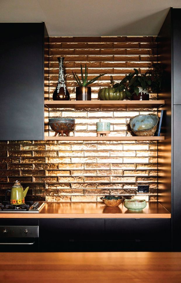 A moody kitchen vignette featuring dark matte cabinetry, warm timber benchtops and open timber shelves, set against a glossy textured brown brick-tile splashback. Decorative ceramics, a sculptural vase, plants and a kettle add character, with warm under-shelf lighting highlighting the textured wall.