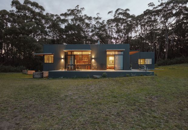 A compact single-storey modern dwelling set within a wooded landscape. The dark corrugated metal-clad facade has several warm-lit glazed openings, a covered terrace or entry zone, and a simple low-profile roof form. The building sits on a broad grassy clearing surrounded by dense eucalyptus-like trees, photographed at dusk.