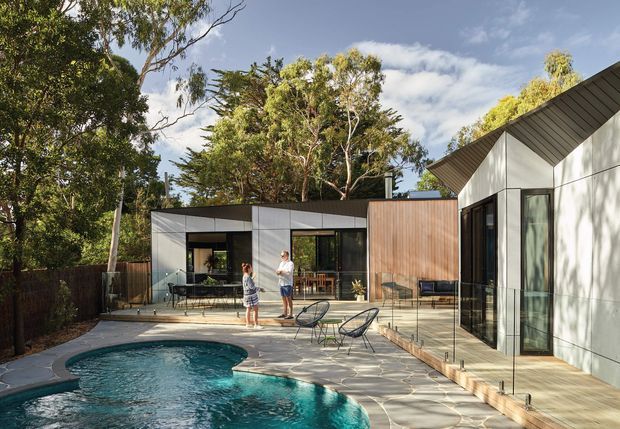 Contemporary residential property viewed from the landscaped pool area, featuring crisp geometric volumes, dark metal-framed glazing, timber-toned wall cladding, pale rendered or panelled walls, and a contrasting steeply pitched roof volume. A paved pool terrace with frameless glass balustrading connects the buildings, with mature trees surrounding the site.