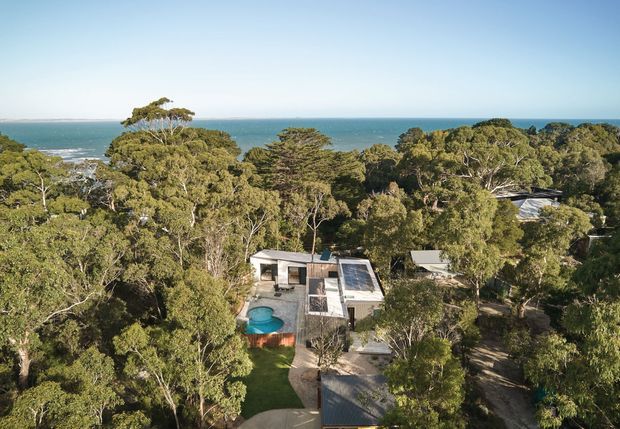Aerial view of a low-rise modern residence nestled among dense coastal trees, with the ocean visible beyond. The home has a flat or low-pitched roof, light-coloured walls, rooftop solar panels, a swimming pool, and a driveway approached through the landscaped bush setting.