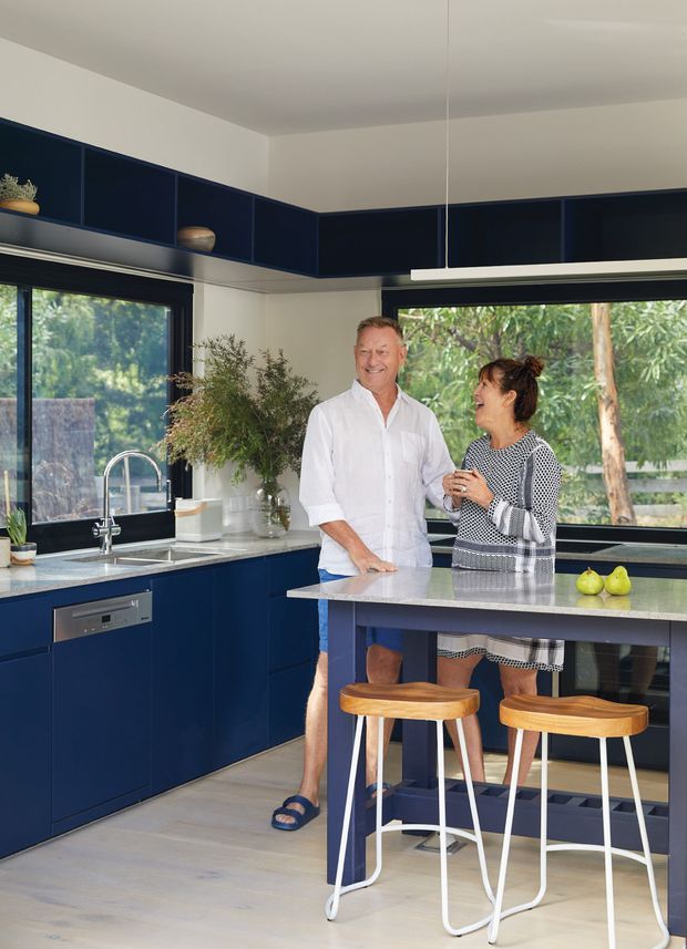 Bright contemporary kitchen with deep navy-blue cabinetry wrapping around two walls and extending above the windows, pale stone benchtops, a central island with integrated seating, stainless-steel sink and dishwasher, and large black-framed windows overlooking greenery. Two people stand at the island, with timber-and-white bar stools and minimal pendant lighting completing the space.