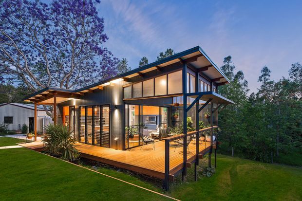 A contemporary elevated timber-and-metal house set among mature trees, photographed at dusk. The two-storey home features extensive glazing, dark structural framing, warm interior lighting, a broad sloping roof, and a raised timber deck supported on posts. Landscaped lawn and dense vegetation surround the building.