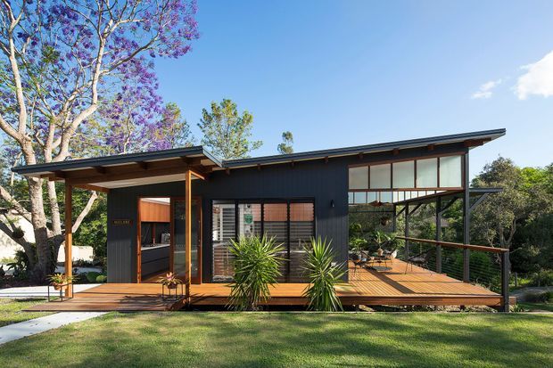 A single-storey contemporary house set within a landscaped garden, with dark vertical cladding, extensive glazing, a shallow skillion roof, and a broad timber deck. The elevated-looking structure features an open-plan living area visible through the glazing, slender timber posts, and a sheltered entry beneath a projecting roof. Bright daylight, lawn, mature trees, and planted yuccas surround the building.