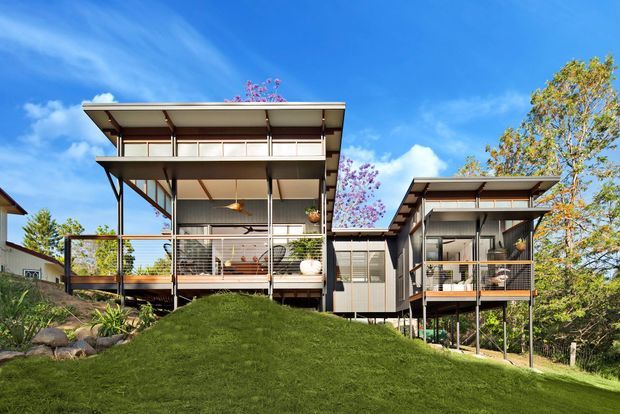 Exterior view of a contemporary elevated residential building on a sloping, landscaped site. Two connected pavilion-like volumes feature broad low-pitched roof planes, slim dark structural posts, extensive glazing, covered balconies, and raised decks. The home is surrounded by lawn, rocks, and mature trees under a bright blue sky.