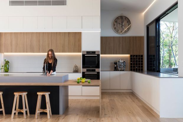 Bright contemporary kitchen with a large island, integrated sink and appliances, white flat-panel cabinetry, warm timber-look joinery, pale timber flooring, and a tiled backsplash. A black-framed glazed opening, wine storage, under-cabinet lighting, and timber bar stools complete the space.