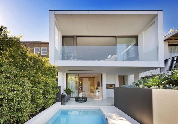 Contemporary two-storey white residence viewed from the front, featuring a projecting upper-level balcony with clear glass balustrade, recessed ground-floor living area, clean rectangular lines, and a narrow blue swimming pool leading toward the entry. Lush planted screening and a dark rendered boundary wall frame the property.