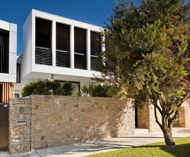 Contemporary two-storey residential facade with crisp white rendered forms, tall dark louvred window screens, and a low natural stacked-stone boundary wall. A mature tree, paved footpath, and lawn occupy the foreground, while a recessed entry gate is visible at the side.