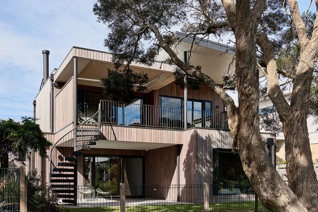 Exterior view of a contemporary multi-level residential house set among mature trees. The home features vertical timber-toned cladding, extensive dark-framed glazing, projecting flat roof planes, a covered upper balcony, and an external spiral staircase. A fenced garden and established vegetation surround the building.
