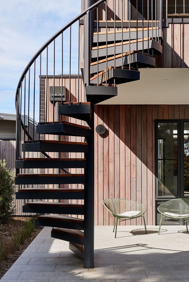 Exterior architectural photograph showing a striking black metal spiral staircase with open treads and slender vertical balustrades beside a modern timber-clad building. The facade combines warm vertical timber boards, dark-framed glazing, and a sheltered concrete-paved terrace with two outdoor wire-frame chairs. Strong sunlight creates crisp shadows, highlighting the staircase and material contrast.