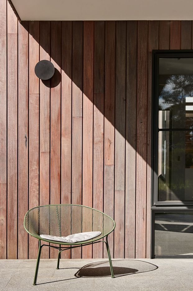 A close-up exterior architectural view featuring vertically arranged dark timber wall cladding, a black-framed glazed door or window, and a green wire lounge chair on a pale concrete terrace. Strong sunlight casts a sharp diagonal shadow across the timber facade, creating a warm, graphic composition.