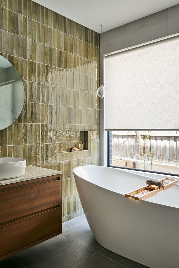 Contemporary bathroom featuring a freestanding white bathtub beside a large window with a roller blind, a timber floating vanity with a white benchtop, and a full-height wall of vertically arranged, weathered green-grey glazed tiles. A round mirror and recessed wall niche are partially visible. The room has a refined, spa-like atmosphere with natural light and muted earthy tones.