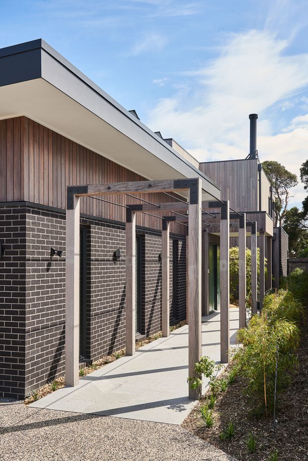 Exterior architectural photograph of a contemporary single-storey building with dark face-brick walls, vertical timber cladding, broad flat roof overhangs, and a covered walkway formed by timber posts and beams. The walkway leads alongside tall narrow windows and landscaped garden beds. The composition highlights the facade, roofline, entry circulation, and carefully detailed outdoor structure under a bright sky.