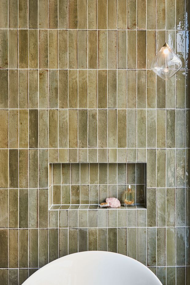 Close-up of a bathroom wall clad in vertically arranged, narrow glazed ceramic tiles in muted olive-green and taupe tones. A recessed tiled niche with a small shelf holds toiletries, while a clear glass pendant light hangs at upper right and the curved edge of a white freestanding bathtub is visible at the bottom. The composition is tactile, refined, and softly lit.