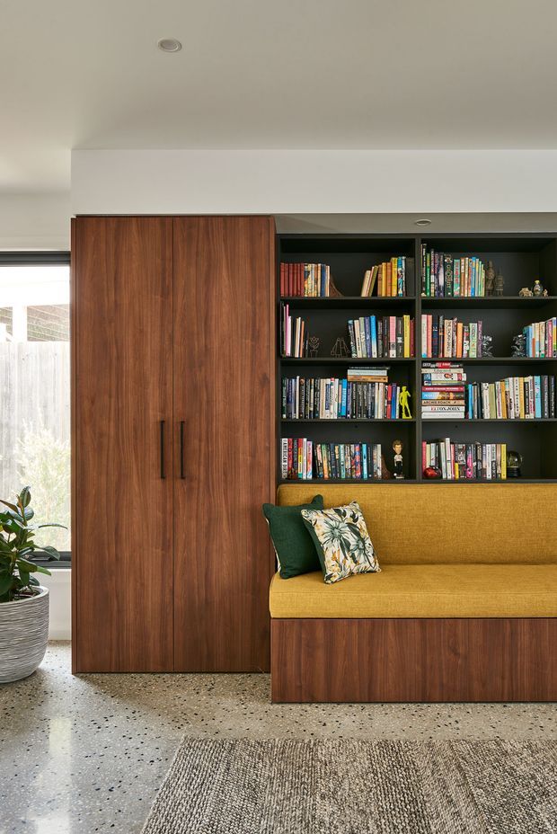Interior view of a contemporary built-in study or living area featuring a tall dark timber storage cabinet, black open bookshelves filled with books, and a mustard upholstered built-in bench seat with timber base. A window, indoor plant, terrazzo-like polished floor, and neutral rug are also visible.