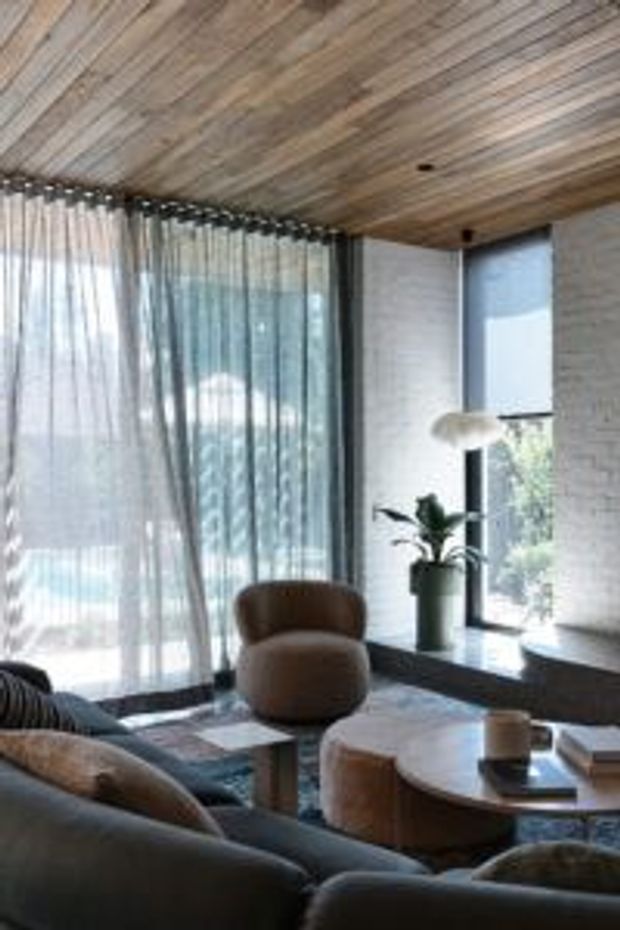 A warm, modern living room with a timber-lined ceiling, pale textured masonry walls, expansive sheer-curtained glazing, and a dark stone or concrete window ledge. Soft rounded upholstered seating surrounds layered low coffee tables, while a potted plant and sculptural pendant add restrained organic character.