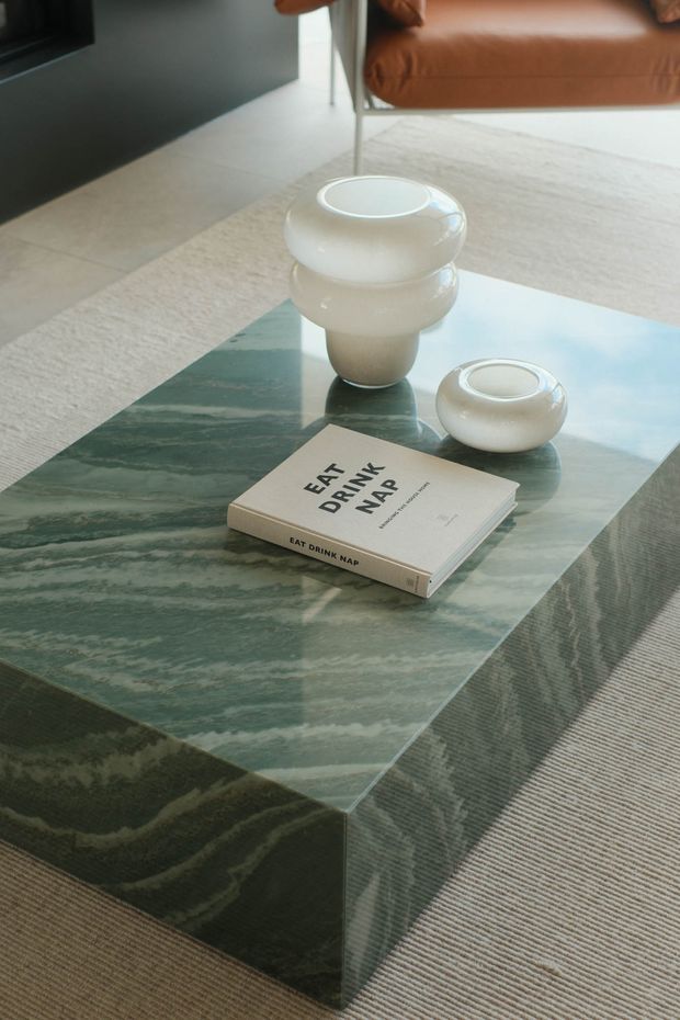A styled living room detail showing a low rectangular coffee table with a glossy green veined stone top and matching waterfall sides. White sculptural ceramic vessels and a design book sit on the table, with a tan leather lounge chair, pale rug, dark wall, and neutral tiled flooring in the background.