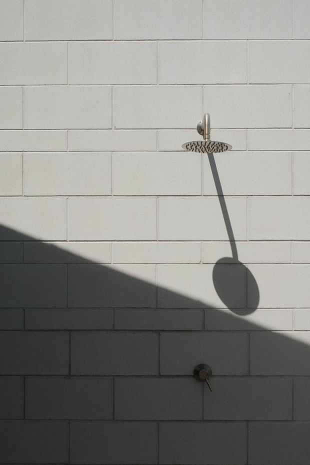 A minimalist shower wall clad in pale rectangular tiles, featuring a round overhead showerhead and separate control fitting. Strong directional sunlight casts pronounced shadows across the tiled surface.