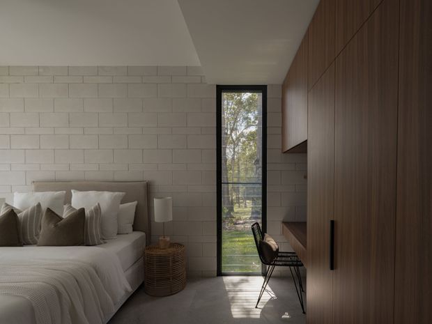 A compact, refined bedroom with a low-profile upholstered bed, layered neutral bedding, a woven bedside table and lamp, exposed pale masonry block walls, polished concrete flooring, and a full-height black-framed window. A built-in dark timber wardrobe and desk occupy the right wall, with a slim black wire chair at the workspace. Natural light and tree views create a calm, warm atmosphere.
