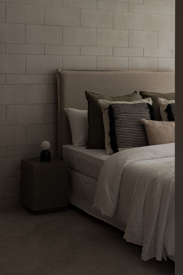 A moody, close-up bedroom interior featuring a large upholstered taupe headboard, layered neutral bedding and pillows, and a simple dark bedside table with a small spherical lamp. The room has a pale grey-beige painted block wall and dark concrete-look flooring, creating a restrained, tactile and contemporary atmosphere.