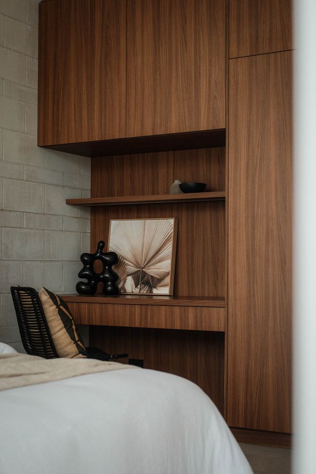 A warm, moody bedroom corner featuring extensive dark timber built-in cabinetry and open shelving, a compact integrated desk, decorative objects, and a partially visible bed with neutral bedding. A grey concrete block wall contrasts with the rich wood joinery, while a dark woven chair and black sculptural decor add texture and visual weight.