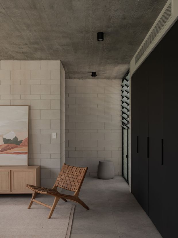 A moody contemporary interior corner featuring exposed concrete ceilings, pale concrete block walls, and a polished concrete floor. A black built-in storage wall with long vertical handles lines the right side, while a woven timber lounge chair, low timber console, neutral rug, ceramic vessel, and abstract artwork add warmth. Narrow louvred glazing and a linear ventilation grille bring filtered light and texture to the space.