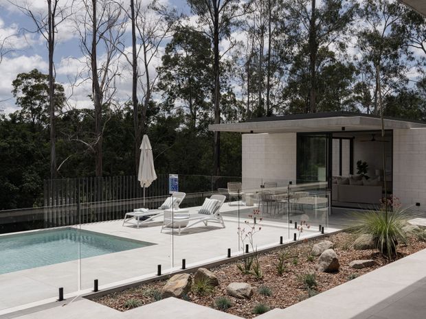 A modern single-storey poolside pavilion or residence with a flat roof, pale masonry walls, extensive glazing, and a broad light-coloured paved terrace. The terrace includes a glass pool fence, swimming pool, outdoor lounge furniture, dining setting, and umbrella. Native trees and a drought-tolerant landscaped garden surround the contemporary outdoor entertaining area.