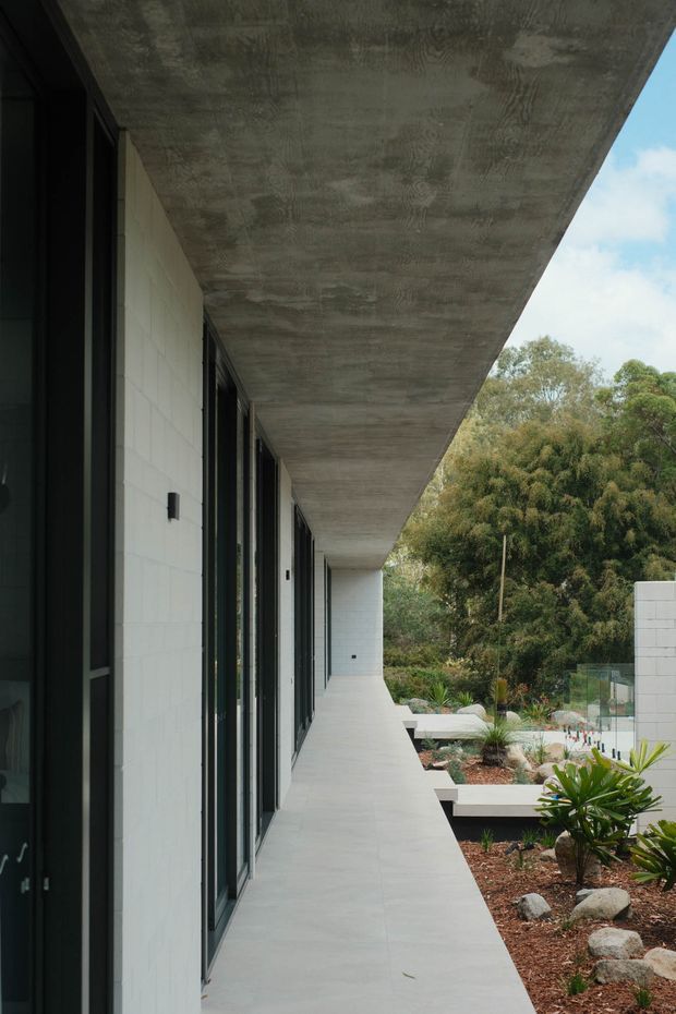 A long, minimalist covered outdoor walkway runs alongside a contemporary building. The space features a broad exposed concrete soffit, pale rectangular masonry wall panels, dark-framed full-height glazing, and a smooth light-grey concrete floor. Landscaped garden beds with rocks and low planting line the open side, with a glass balustrade and trees beyond.