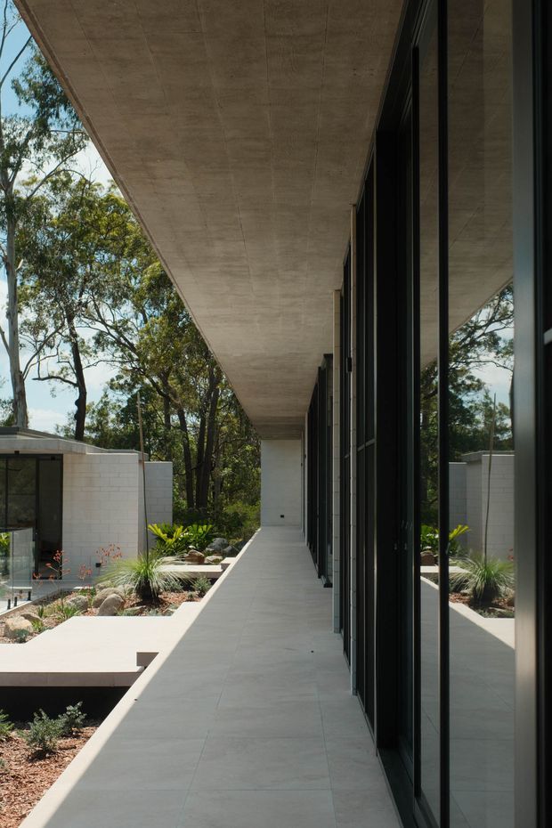 A long covered exterior walkway beside a modern building, with a broad exposed concrete canopy, dark-framed floor-to-ceiling glazing, pale tiled paving, and landscaped courtyards with rocks and native planting. The glazing reflects the surrounding trees and garden, reinforcing the connection between the building and its wooded site.