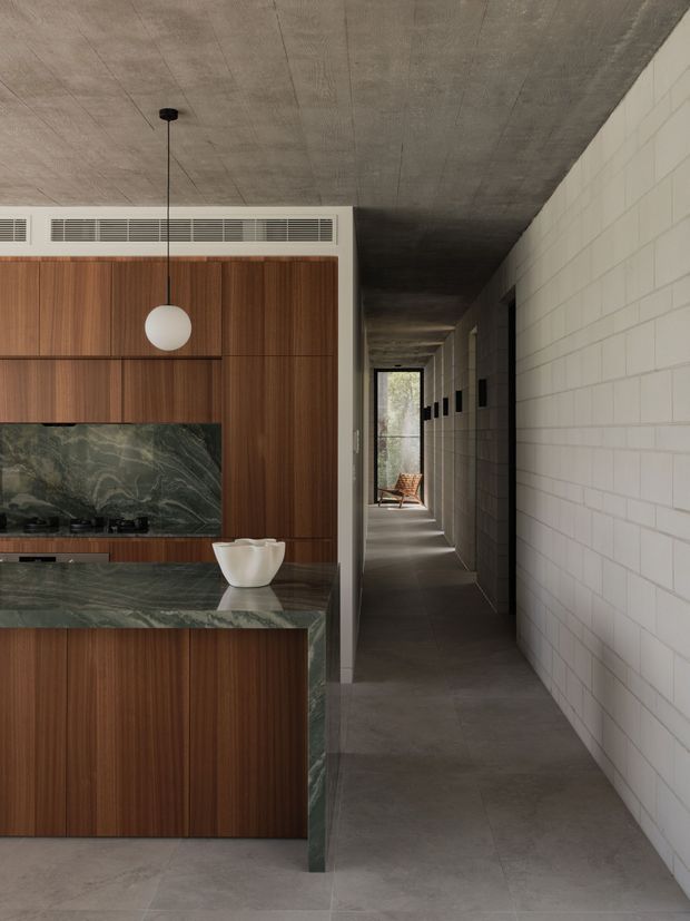 A refined contemporary kitchen viewed from the main living area, with warm vertical timber cabinetry, a dark green veined stone backsplash and waterfall island, and a simple spherical pendant light. The kitchen opens onto a long, dimly lit corridor lined with pale rectangular masonry blocks and dark concrete surfaces. The space has strong linear geometry, integrated ventilation, and a restrained material palette.