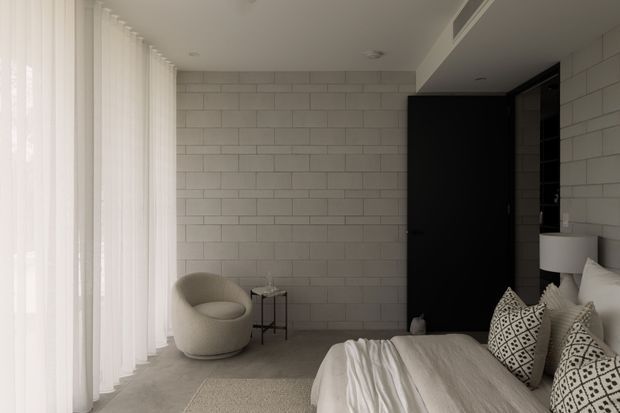 A serene contemporary bedroom with a bed in the foreground, a curved upholstered lounge chair, small side table, and tall sheer curtains along the left wall. The room features a pale grey block-pattern tiled or masonry feature wall, dark built-in wardrobe or door panels, recessed ceiling lighting, and a restrained neutral palette.