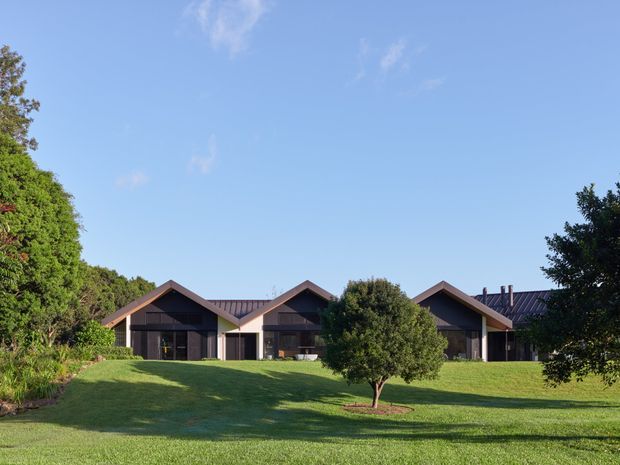 A low-rise contemporary residential building set within a broad manicured lawn and mature landscaped garden. The facade is composed of several connected pitched gable-roof forms with dark metal roofing, deep roof overhangs, dark wall cladding, and large glazed openings. The image is photographed from the landscaped grounds under a clear blue sky.
