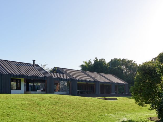 A low, elongated contemporary residence set within a broad grassy landscape. The building has dark vertical cladding, standing-seam metal gabled roofs, extensive glazing, and covered outdoor living areas. Mature trees surround the home, with a bright, open setting and strong horizontal composition.