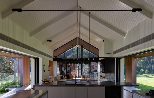A spacious open-plan kitchen and living area with a large central island, dark stone benchtop, and extensive floor-to-ceiling glazing. The room features a dramatic pitched, white-lined ceiling with exposed timber structural beams, black linear pendant lighting, timber wall and window detailing, and a glazed gable end overlooking landscaped grounds. The interior has a refined contemporary character with strong natural light and indoor-outdoor connection.