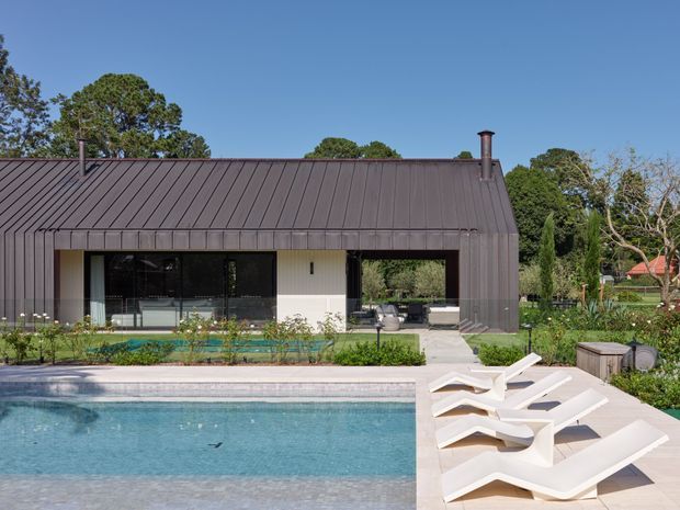 A contemporary single-storey pavilion-style house viewed from a swimming pool. The building has a dark standing-seam metal pitched roof and matching vertical wall cladding, expansive black-framed glazing, and a covered outdoor living area. The foreground features a rectangular pool, pale paved decking, white sculptural sun loungers, and landscaped planting.