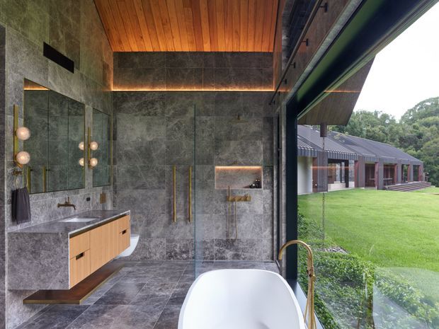 A luxurious contemporary bathroom with a freestanding white bathtub positioned beside full-height glazing overlooking a landscaped lawn and pavilion-like building. The room features large grey stone-look wall and floor tiles, a timber vanity with integrated basin, tall mirrored cabinetry, brass-toned fixtures, a frameless glass shower enclosure, concealed warm LED strip lighting, and a slatted timber ceiling. The open connection to the exterior creates a bright, refined indoor-outdoor atmosphere.