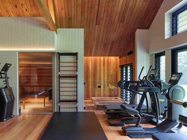 A sophisticated home gym with multiple cardio machines, a black exercise mat, wall-mounted climbing bars, and a glass-fronted sauna. The room features warm timber flooring, extensive timber ceiling lining with exposed beams, vertical timber wall cladding, white ribbed wall panels, and large louvred windows providing natural light.