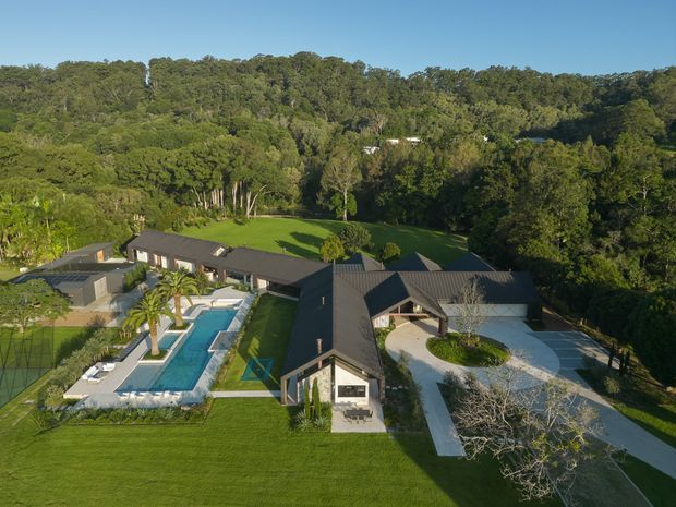 Aerial view of a sprawling contemporary residential property set within a heavily wooded landscape. The dark-roofed, low-rise home is arranged around landscaped lawns and includes a long swimming pool, paved pool terrace, curved driveway, and detached or adjoining outbuildings. Dense forest surrounds the site.