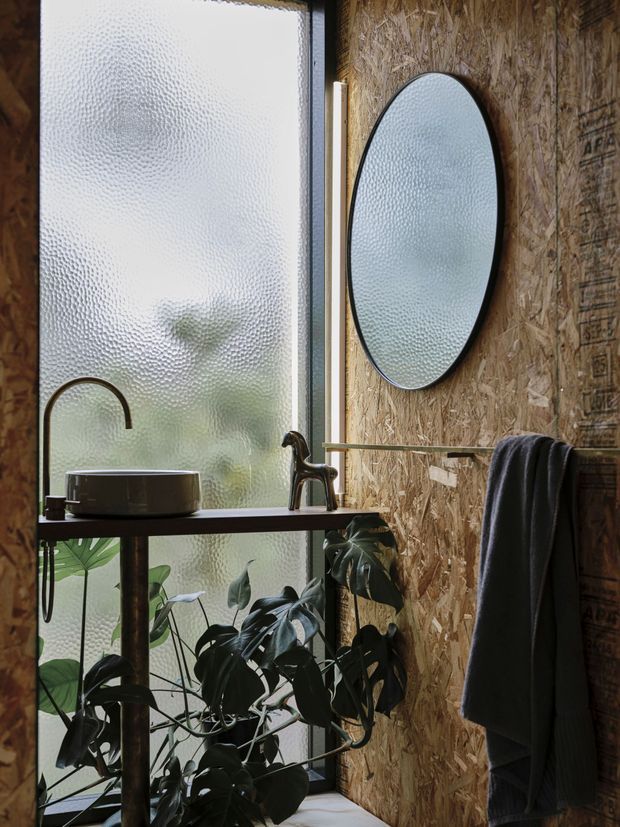 A compact industrial-style bathroom under construction, featuring a black-framed round mirror, wall-mounted towel rail, dark vessel basin with exposed plumbing on a slim metal console, and a dark faucet. The walls are clad in visible oriented strand board, while a large textured translucent window provides diffused natural light. Lush indoor plants are visible below the vanity.