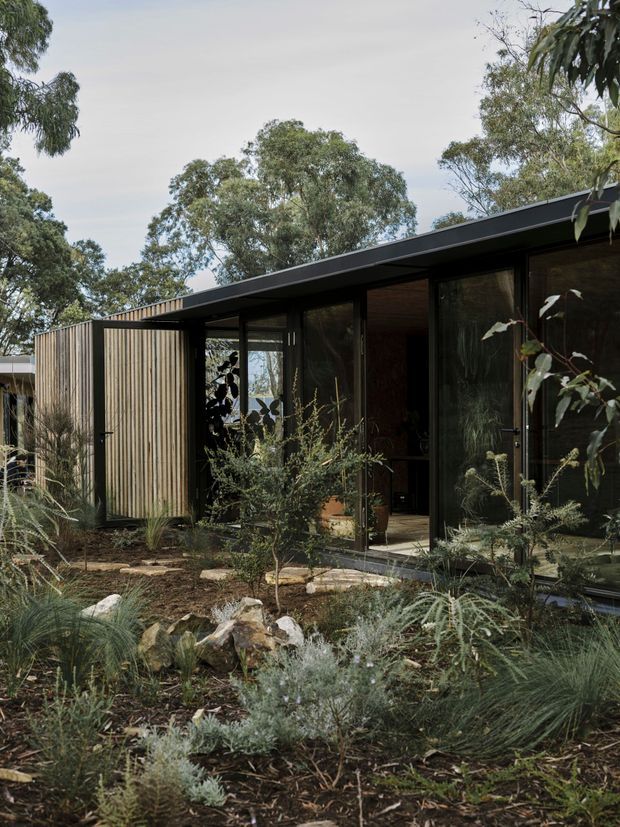 Exterior view of a low, single-storey modern dwelling set within a natural landscaped garden. The building features a long dark-framed glazed facade, a broad shallow roof overhang, and vertical timber screening or cladding at the side. Native-style planting, rocks, grasses, and established trees surround the structure.