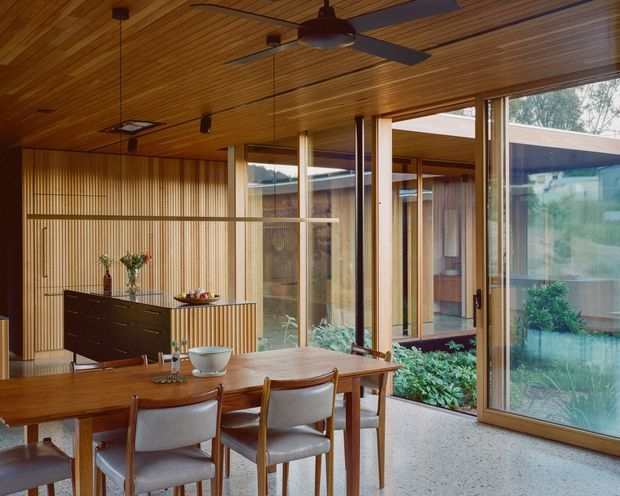 A warm mid-century-inspired dining room with a long timber dining table and upholstered wooden chairs, set against extensive vertical timber wall panelling and a dark built-in sideboard. The room has a timber-lined ceiling with ceiling fans and recessed lighting, polished terrazzo flooring, and large timber-framed glazed doors and windows opening visually onto a planted courtyard. The interior is filled with natural light and has a strong connection to the garden.