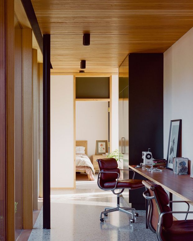 A refined home office viewed along a corridor, with a long dark wood desk, two burgundy leather office chairs, a vintage sewing machine, framed artwork, and open shelving. The office connects visually to a bedroom beyond. Warm timber slat ceilings and wall panelling contrast with white walls, dark vertical joinery, and a polished speckled floor. Slim cylindrical pendant lights and strong linear geometry create a calm, contemporary interior.