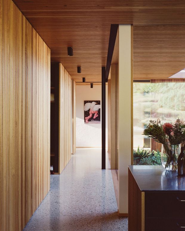 A long, refined residential hallway with vertical timber wall panelling, a matching timber-lined ceiling, dark recessed linear lighting, and pale terrazzo flooring. Full-height glazing on the right overlooks planted landscaping, while a dark built-in console or cabinetry with a stone-like top occupies the foreground. The corridor leads toward an artwork at the far end and has a warm, highly considered material palette.