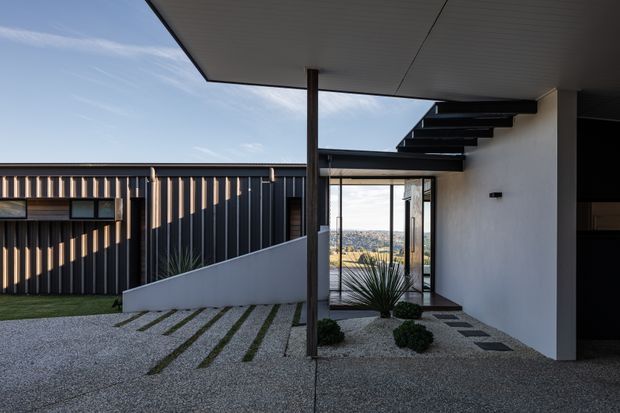 Modern residential entry courtyard viewed from outdoors, featuring a flat-roofed covered porch, dark vertical metal cladding, white rendered walls, exposed steel roof framing, floor-to-ceiling glazing, and a minimalist gravel-and-paving landscape. A distant urban landscape is visible through the entry glazing.